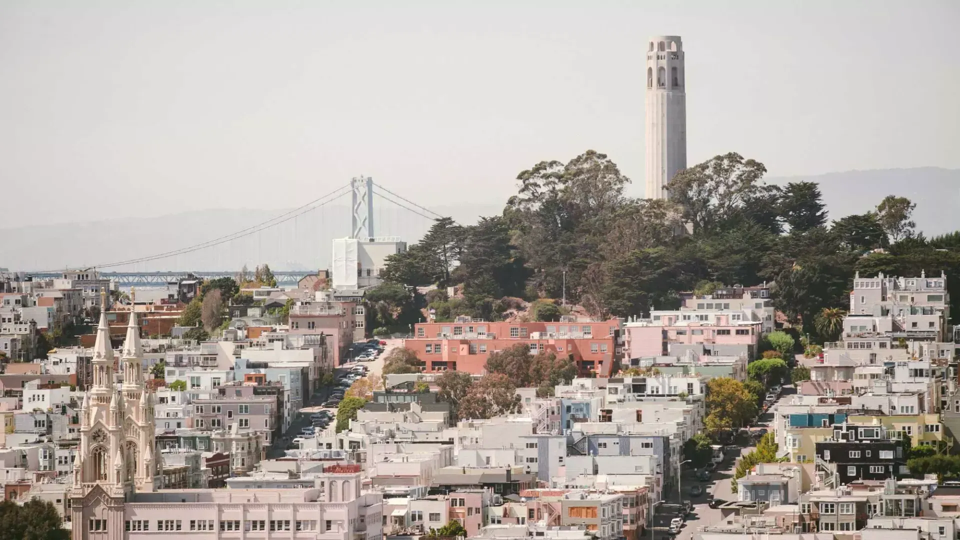 샌프란시스코의 比特塔는 배경에 베이 브릿지가 있고 전경에 집들로 뒤덮인 언덕이 있는 사진입니다.
